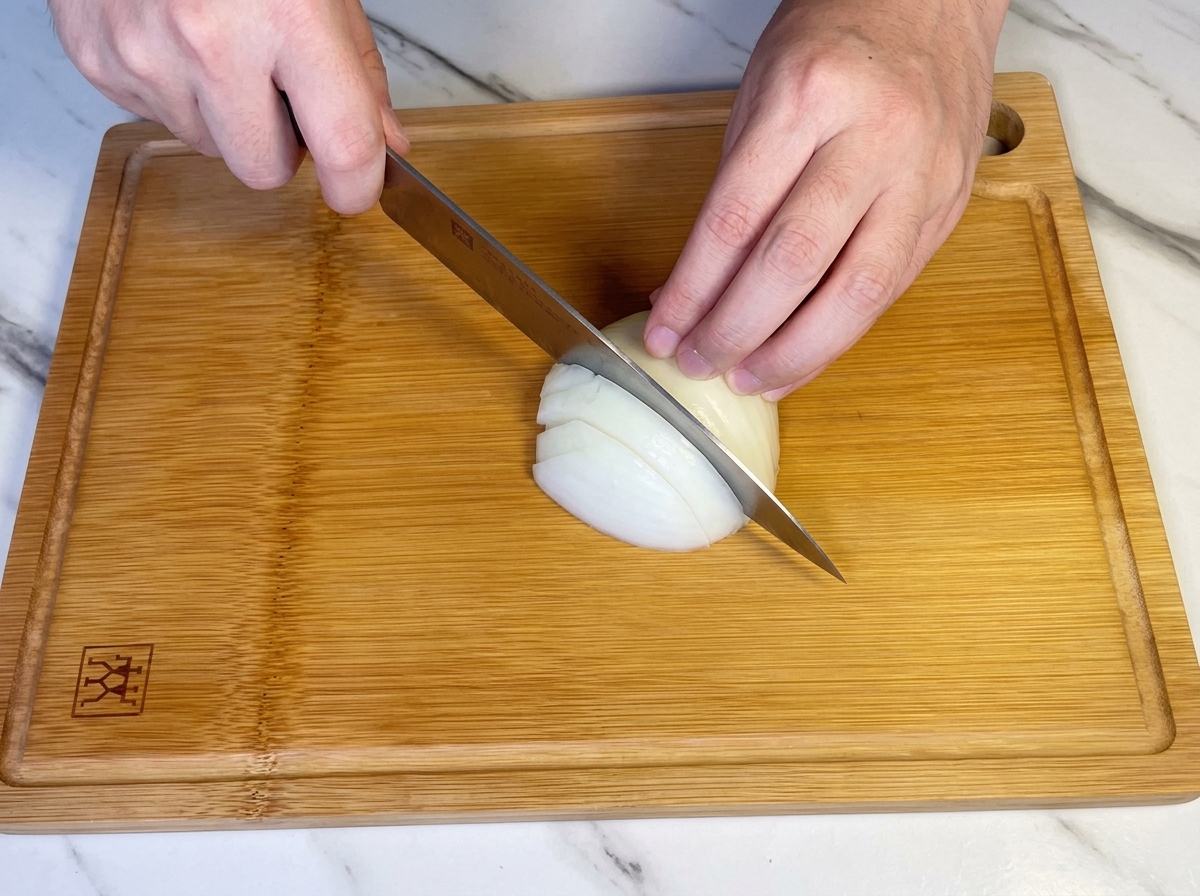 Manos usando un cuchillo de chef para picar media cebolla blanca en una tabla de madera.