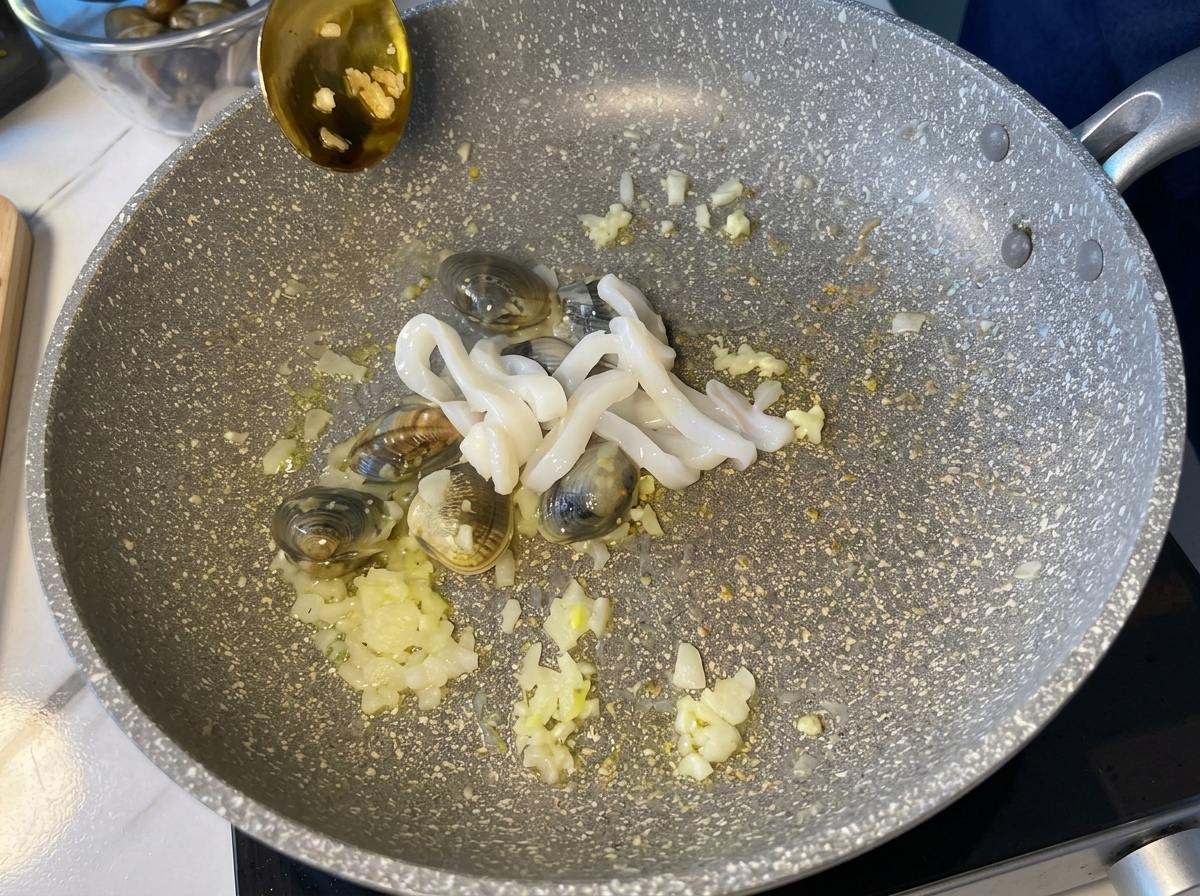 Anillos de calamar blanco crudo siendo añadidos a una sartén con almejas y aromáticos.