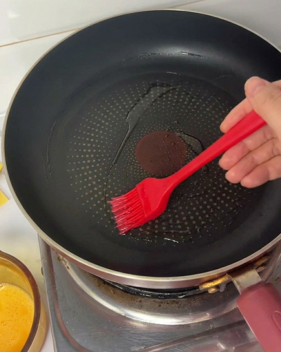 Una mano usando un pincel de silicona rojo para esparcir aceite en una sartén antiadherente negra.