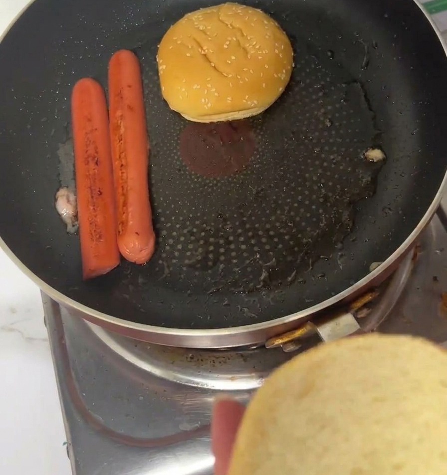 Panes de hamburguesa con sésamo tostándose boca abajo en una sartén junto a las salchichas.