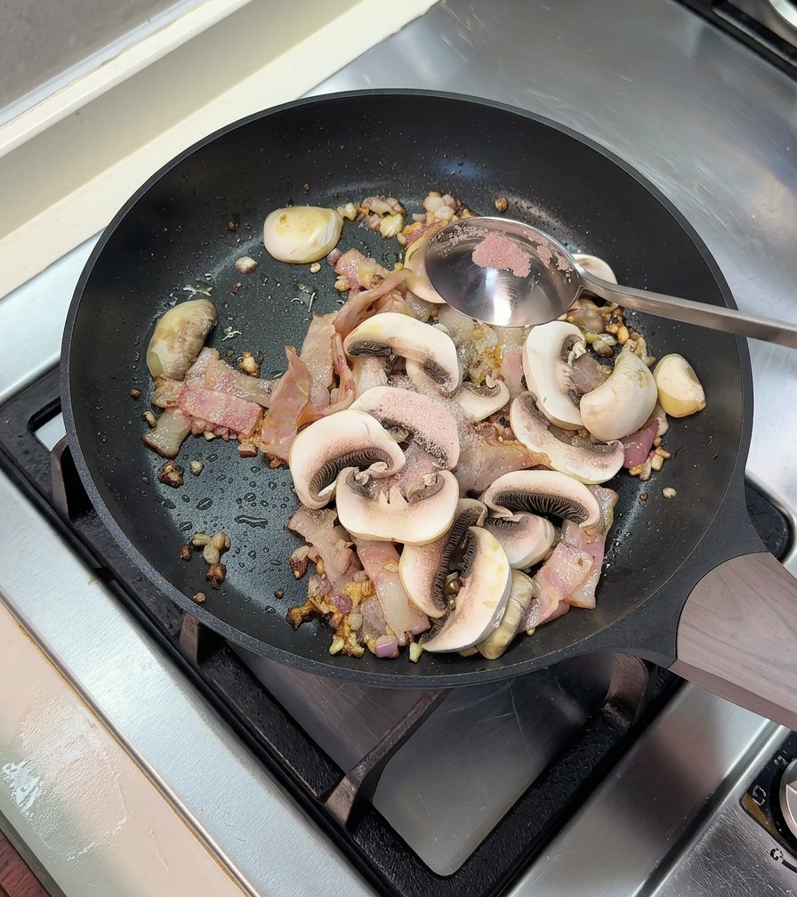 Añadiendo una cucharadita de sal a la sartén con bacon y champiñones cocinados.