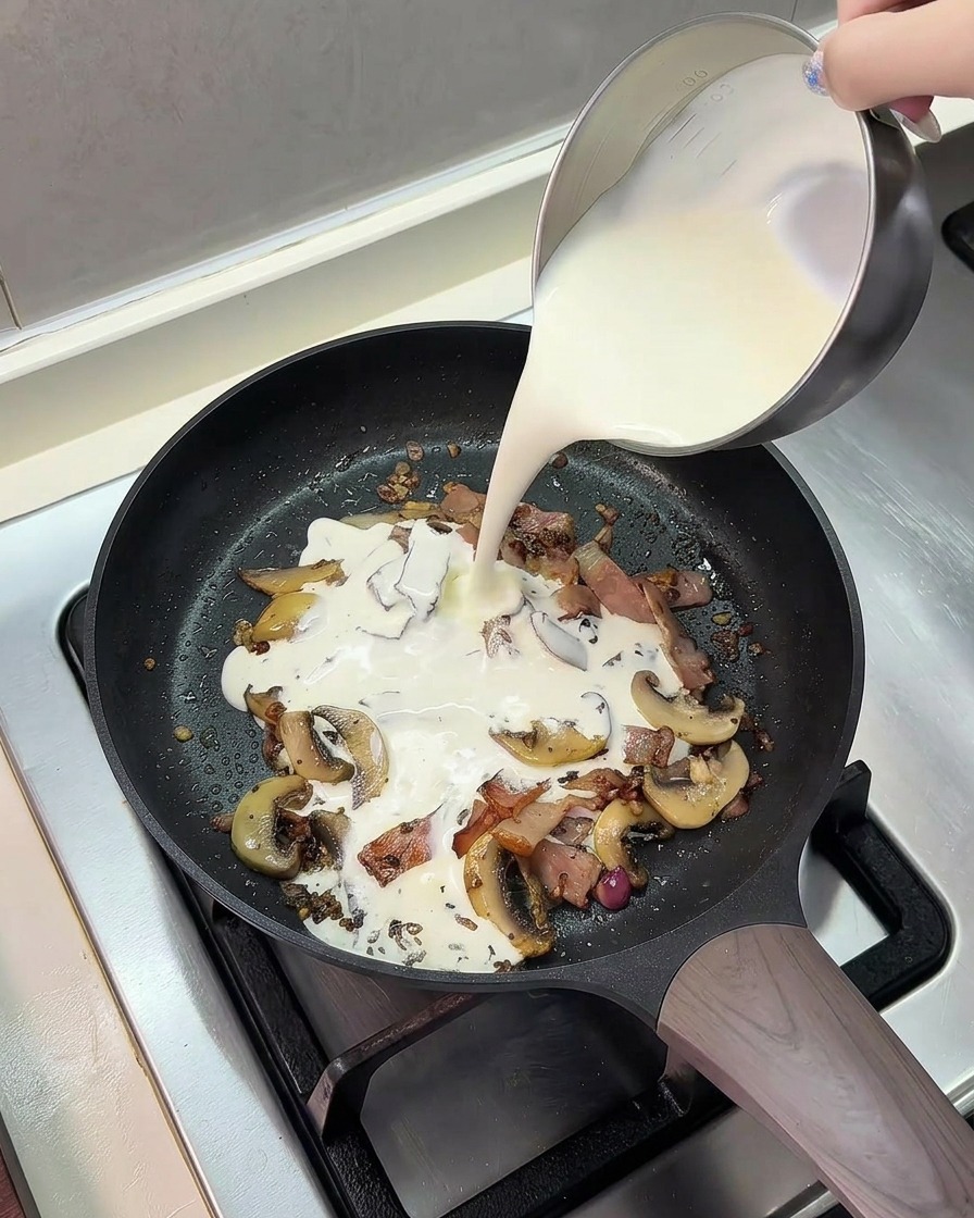 Vertiendo una mezcla de leche y crema de leche desde una jarra metálica a la sartén.