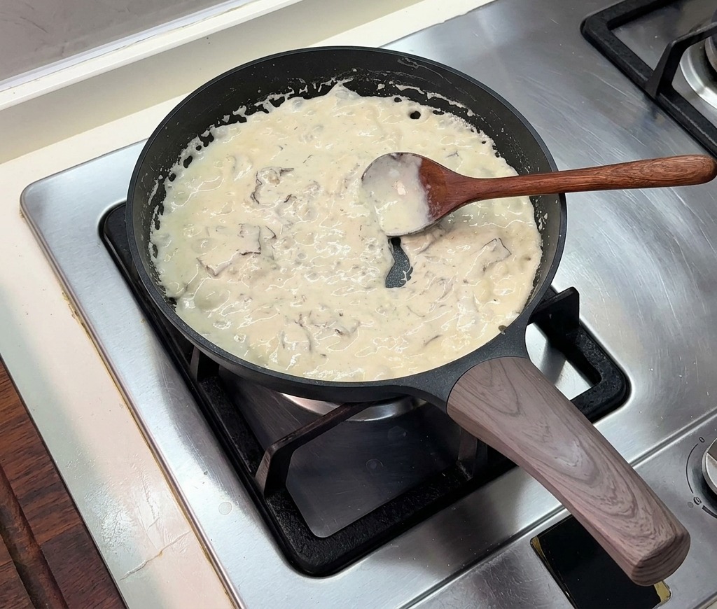 Una salsa de crema espesa burbujeando en la sartén mientras se remueve con una cuchara de madera.