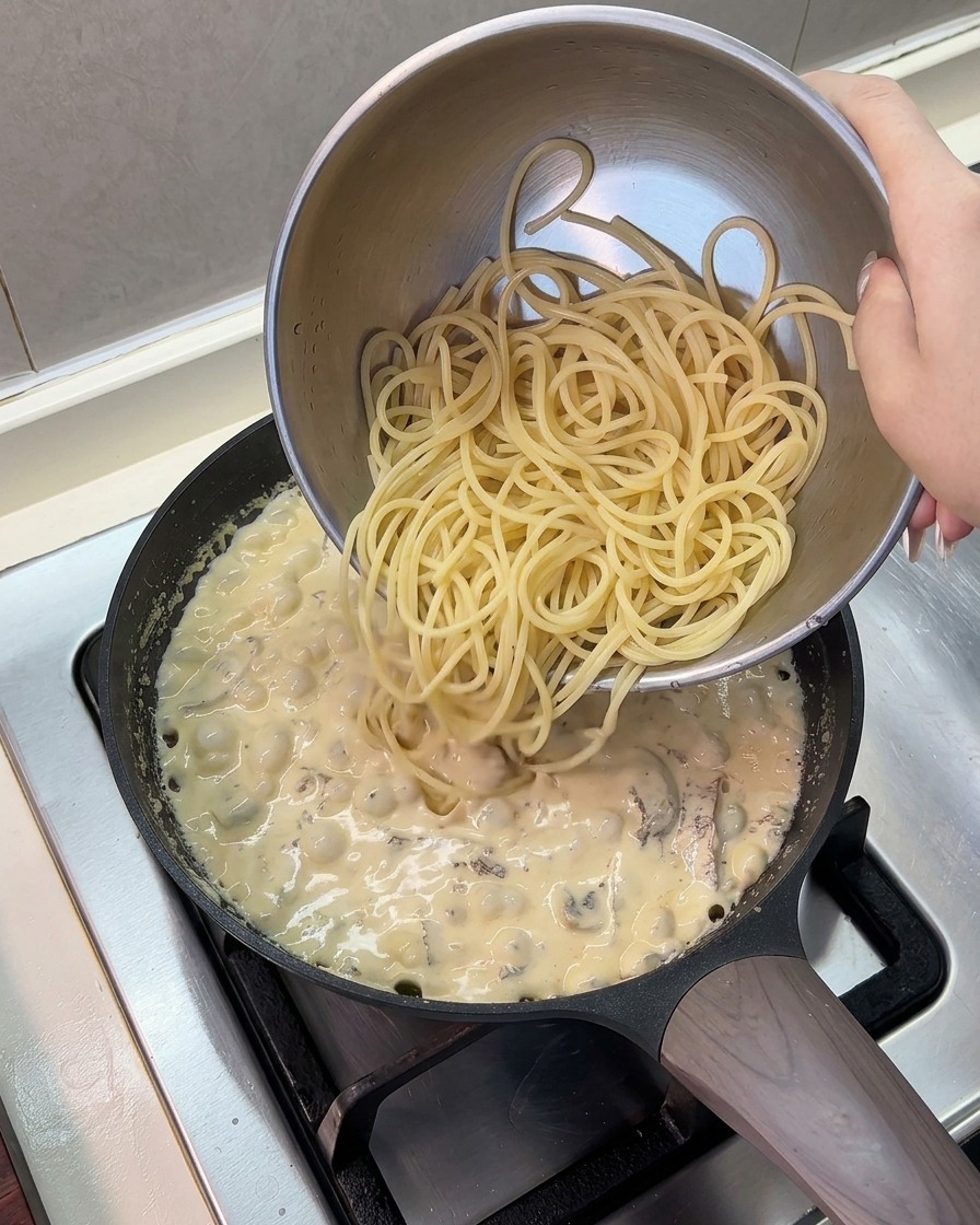 Espaguetis cocidos cayendo desde un bol de acero inoxidable a la sartén con salsa cremosa.