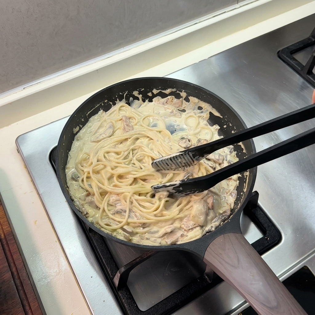 Pinzas de cocina mezclando espaguetis en una rica salsa de crema dentro de la sartén.