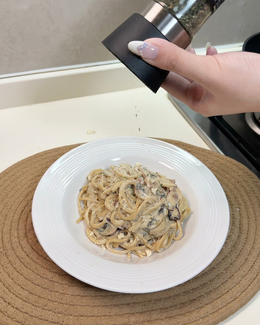 Una mano espolvoreando queso y hierbas sobre un plato caliente de espaguetis cremosos.