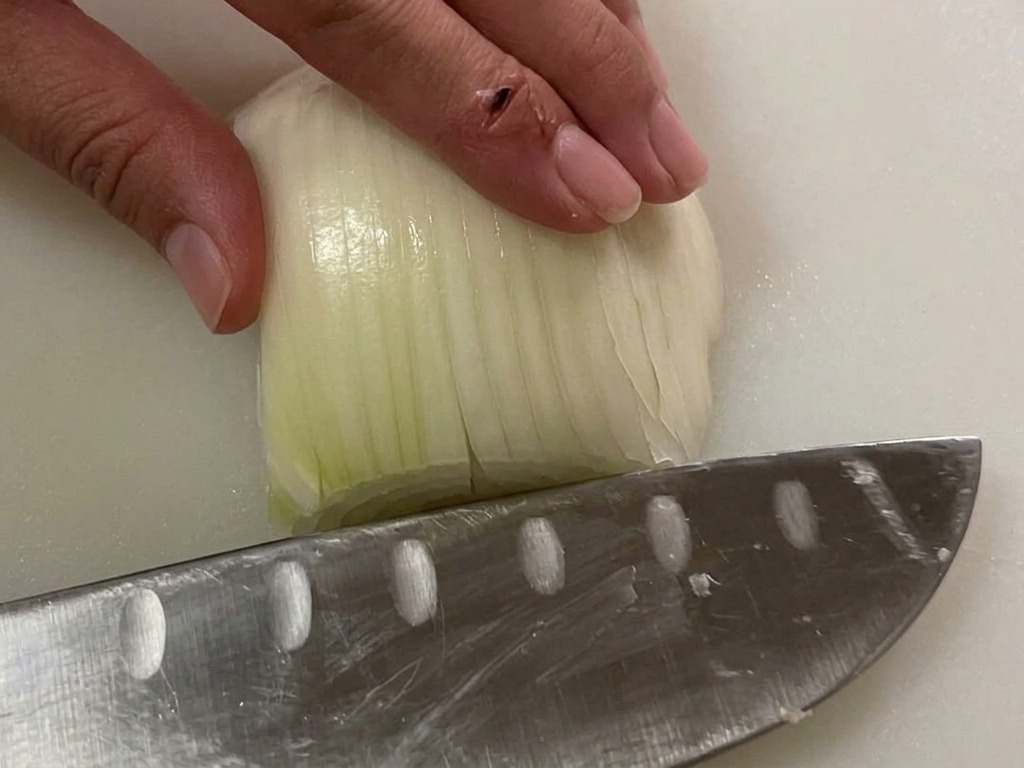 Mano sujetando media cebolla blanca sobre una tabla de cortar mientras un cuchillo se prepara para picarla.