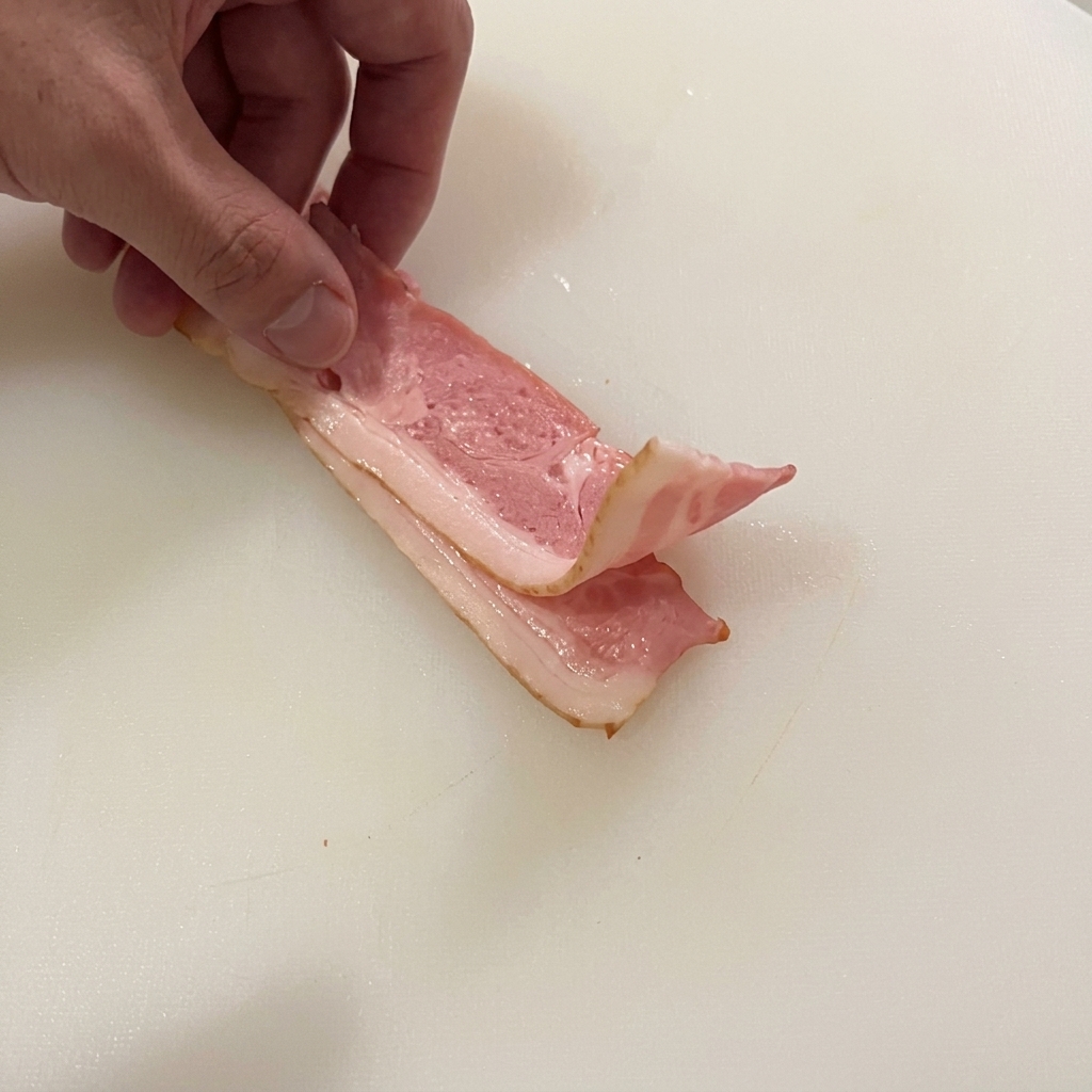 Mano doblando dos tiras de bacon crudo en una tabla blanca, listas para ser cortadas.