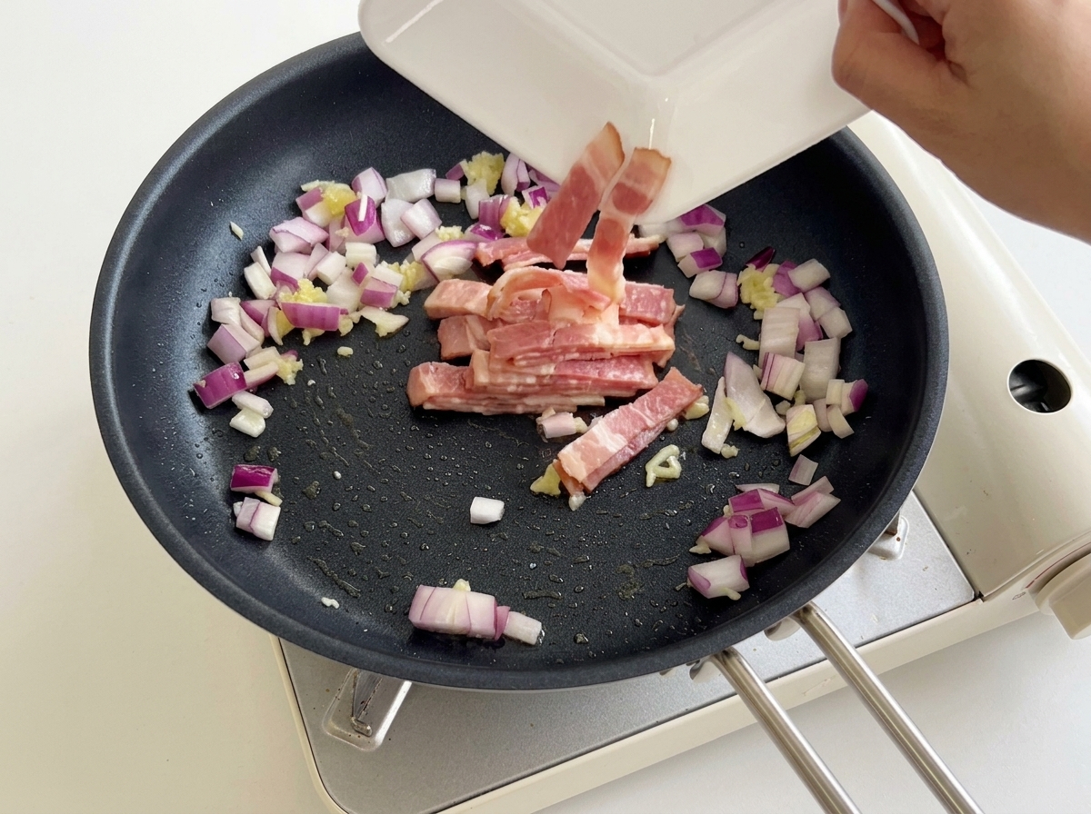 Bacón troceado cayendo desde un plato blanco a una sartén con cebolla roja y ajo.