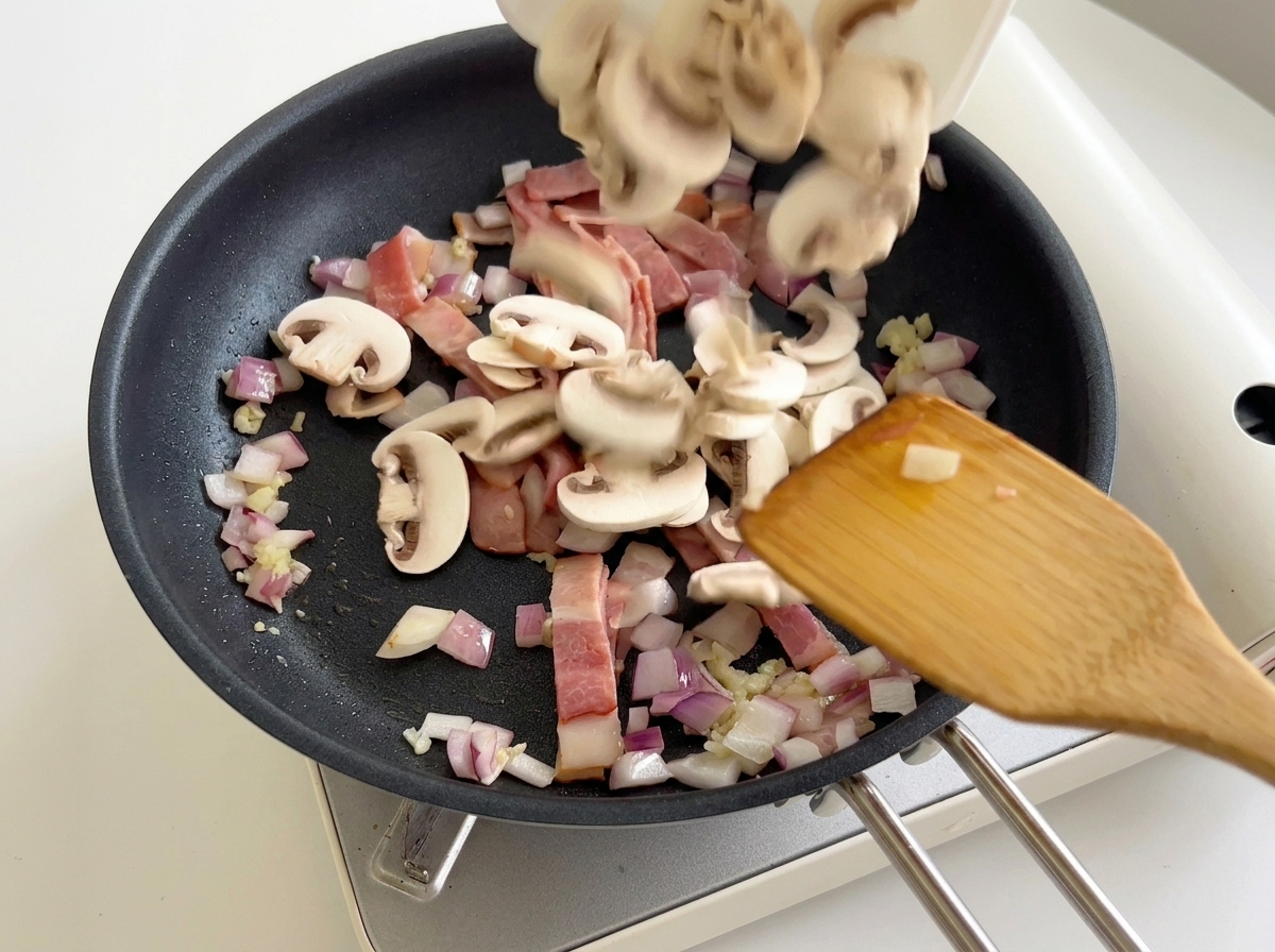 Champiñones laminados siendo añadidos desde un plato a una sartén que contiene bacón y cebollas.