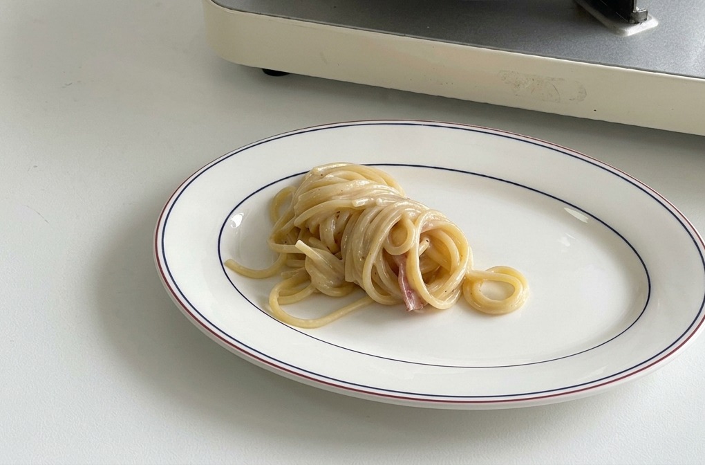 Un nido de espaguetis cremosos perfectamente enrollado en el centro de un plato blanco ovalado con borde azul oscuro y rojo.