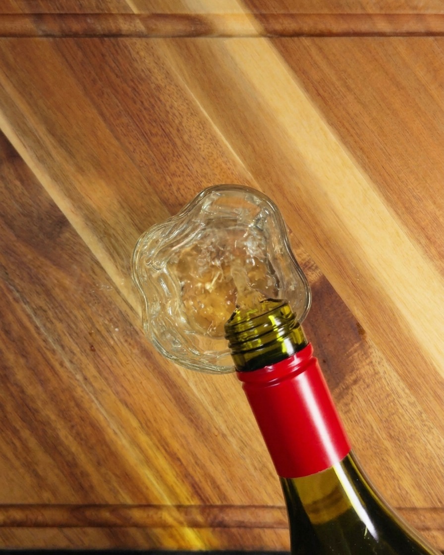 Vino blanco vertiéndose de una botella de vidrio verde en un pequeño vaso transparente sobre una superficie de madera.