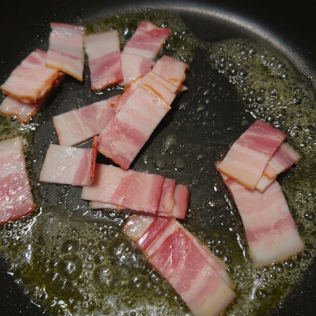 Pequeñas tiras de bacón chisporroteando y dorándose en una sartén con su propia grasa caliente.