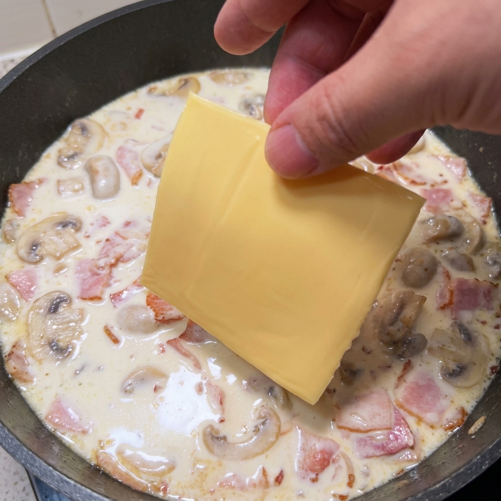 Una mano colocando una loncha de queso amarillo en la sartén con la salsa cremosa.