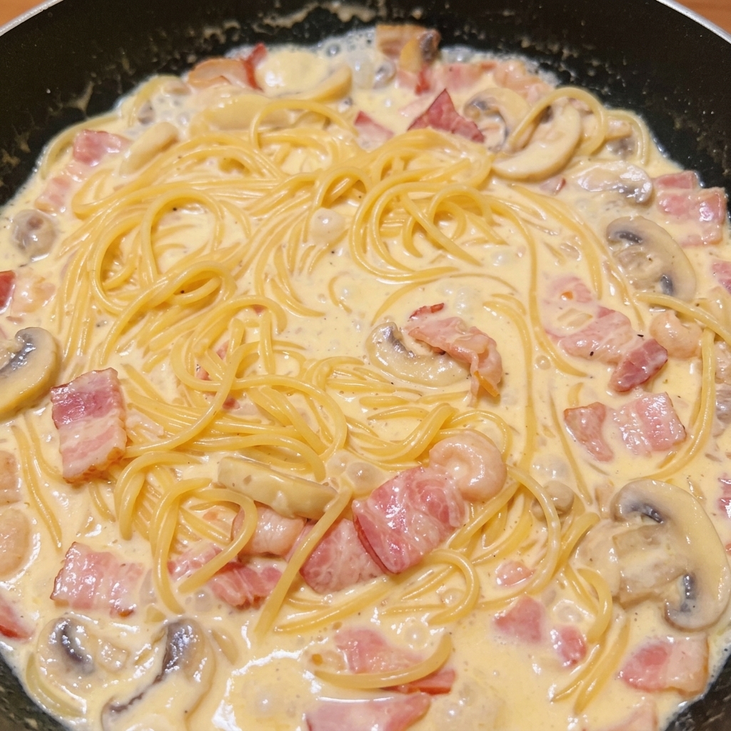 Espaguetis cocidos mezclados con la salsa rica y cremosa de bacón y champiñones en una sartén oscura.