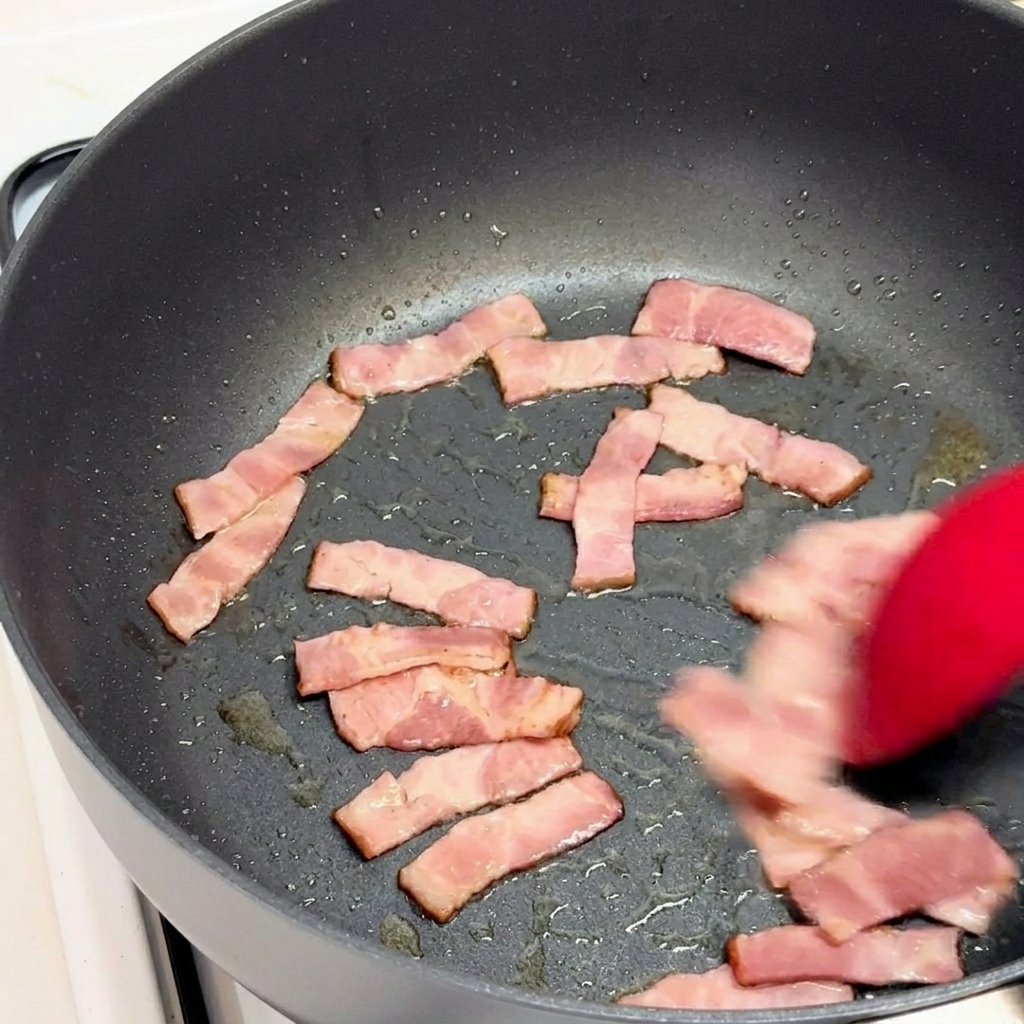Trozos de bacon salteándose con una espátula de silicona roja en una sartén negra.