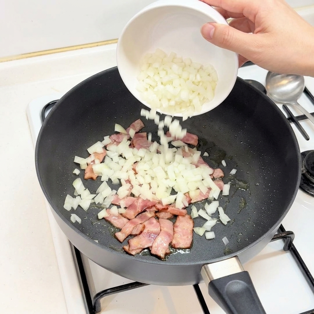 Una mano vertiendo cebolla picada desde un bol blanco a una sartén con bacon cocinado.