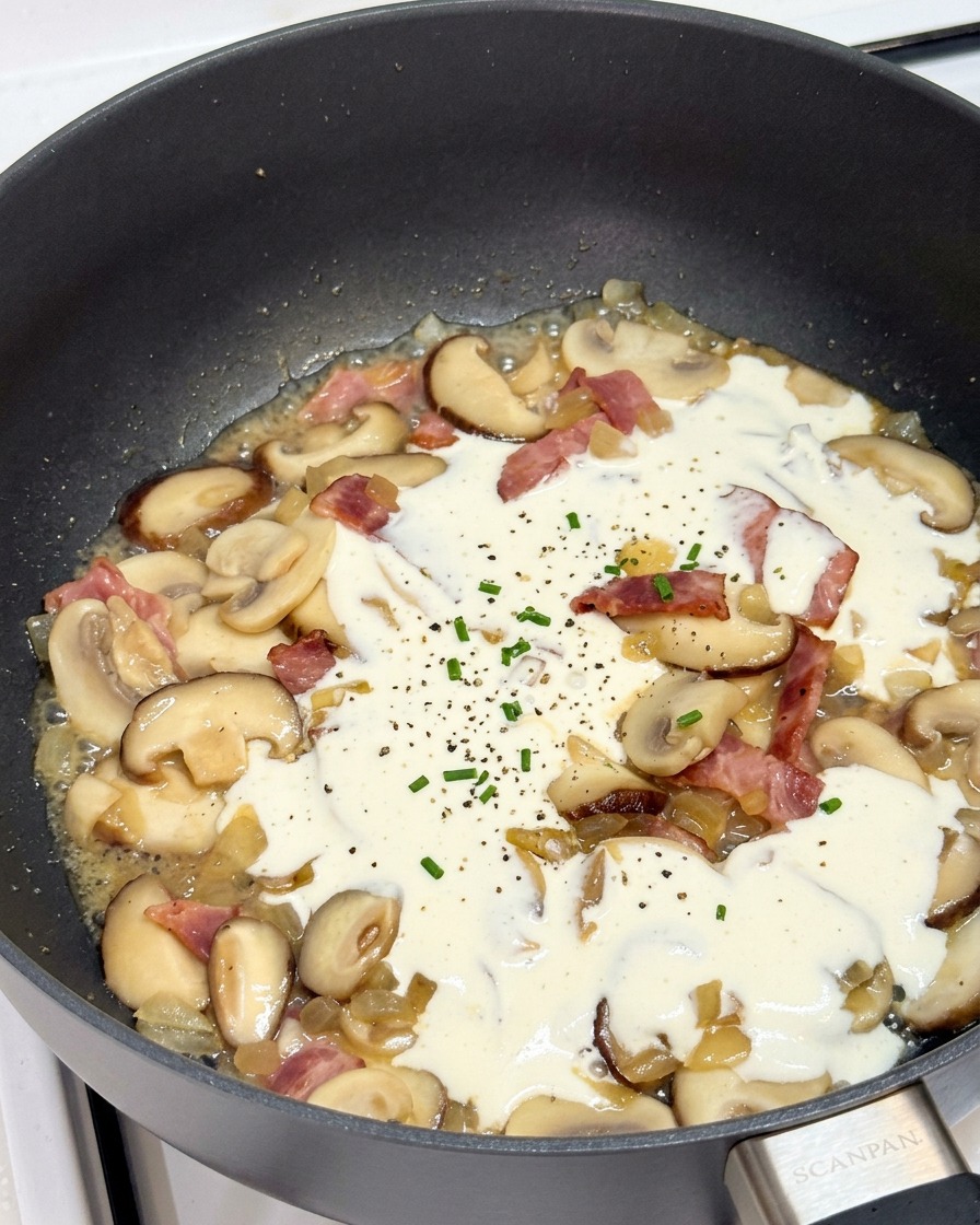 Crema de leche hirviendo a fuego lento en una sartén con champiñones y bacon.