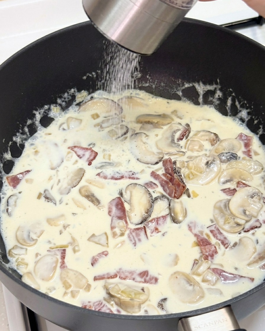 Una persona usando un molinillo para añadir pimienta negra fresca a una sartén con salsa cremosa de setas y bacon.