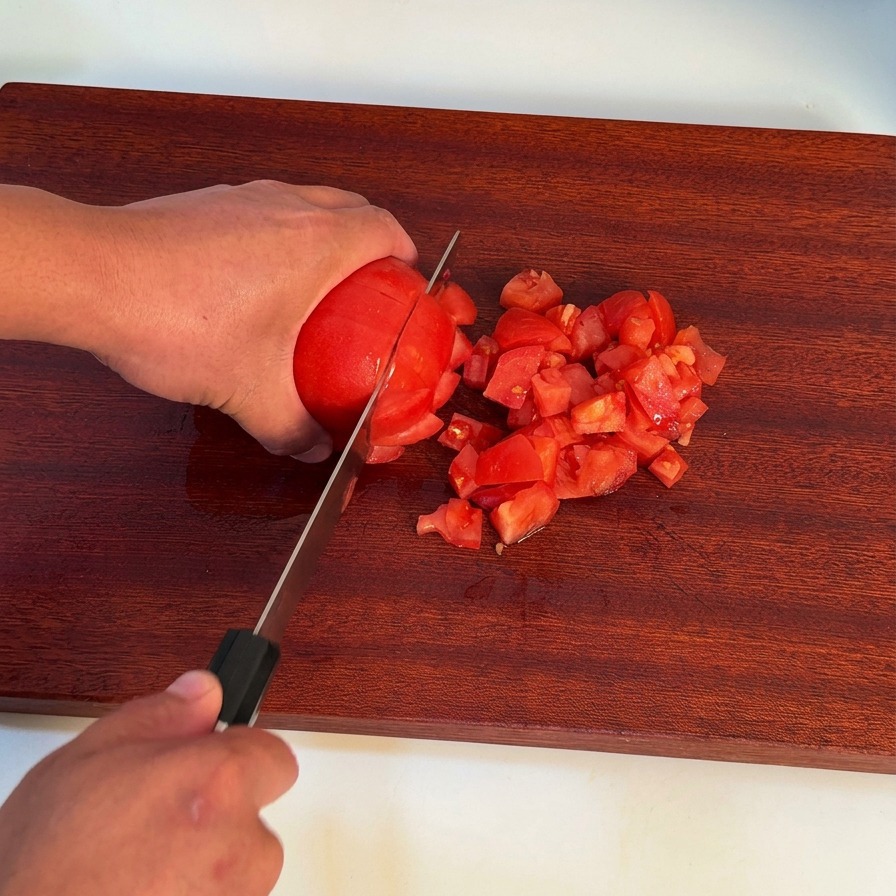 Una mano usando un cuchillo de chef para picar un tomate rojo pelado sobre una tabla de cortar de madera.