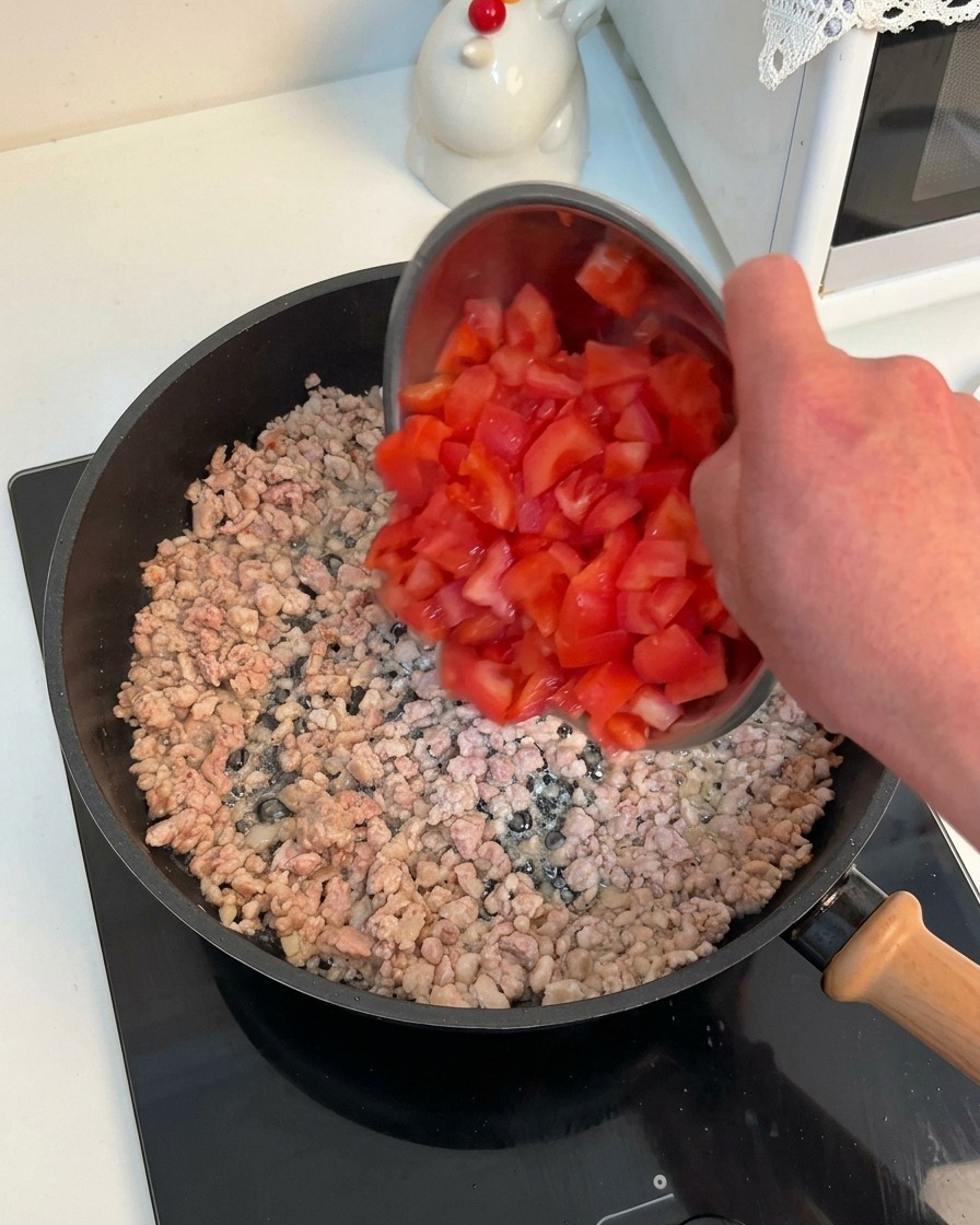 Una mano vertiendo tomates picados en la sartén con la carne dorada.