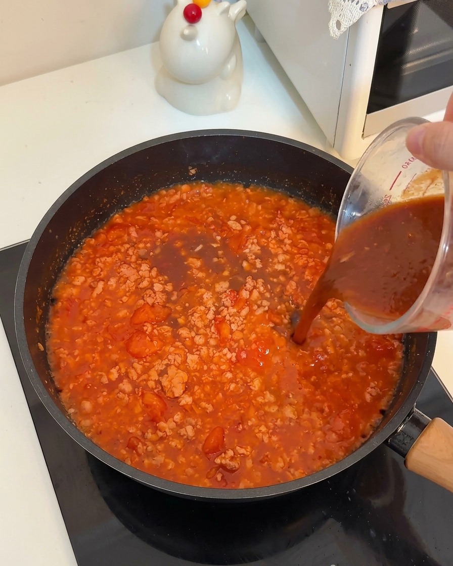 Un líquido oscuro vertiéndose desde una jarra medidora a la sartén con carne y tomates.