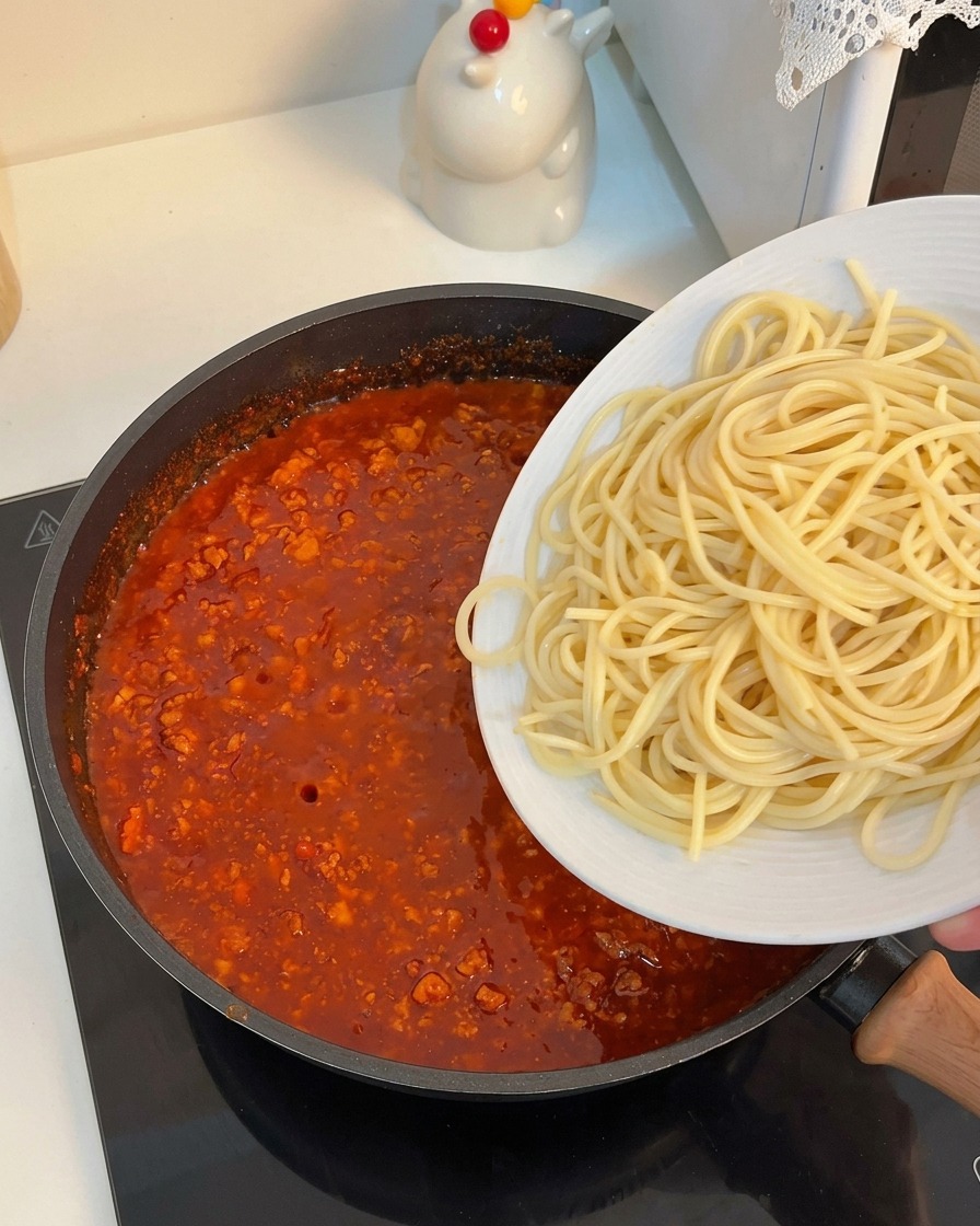 Espaguetis cocidos deslizándose hacia la sartén llena de salsa de carne espesa.