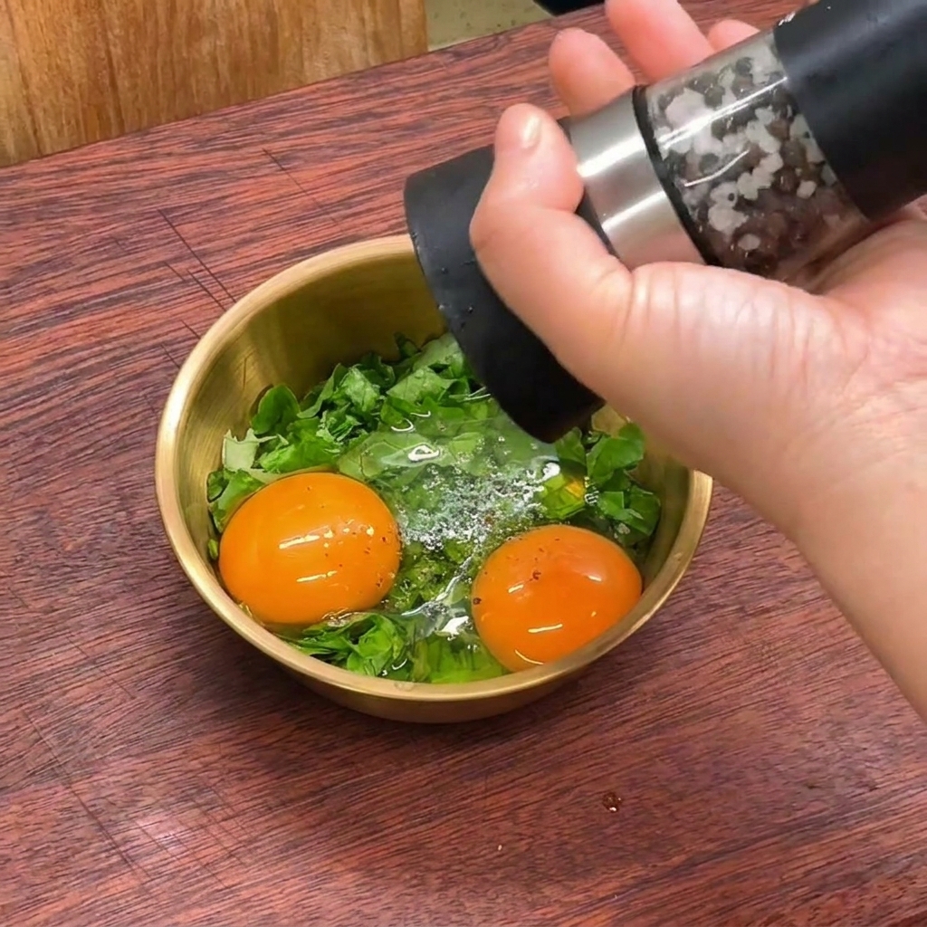 Una mano usando un molinillo de pimienta para moler pimienta negra fresca sobre un bol dorado con huevos crudos y lechuga picada.