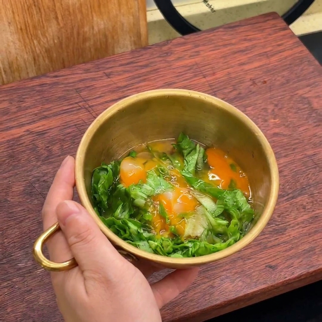 Una mano sosteniendo un bol dorado con lechuga picada y dos huevos crudos recién abiertos.