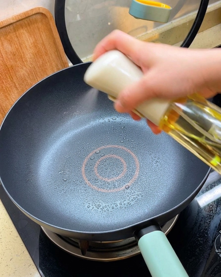 Una mano pulverizando una fina capa de aceite de cocina en una sartén negra antiadherente caliente.