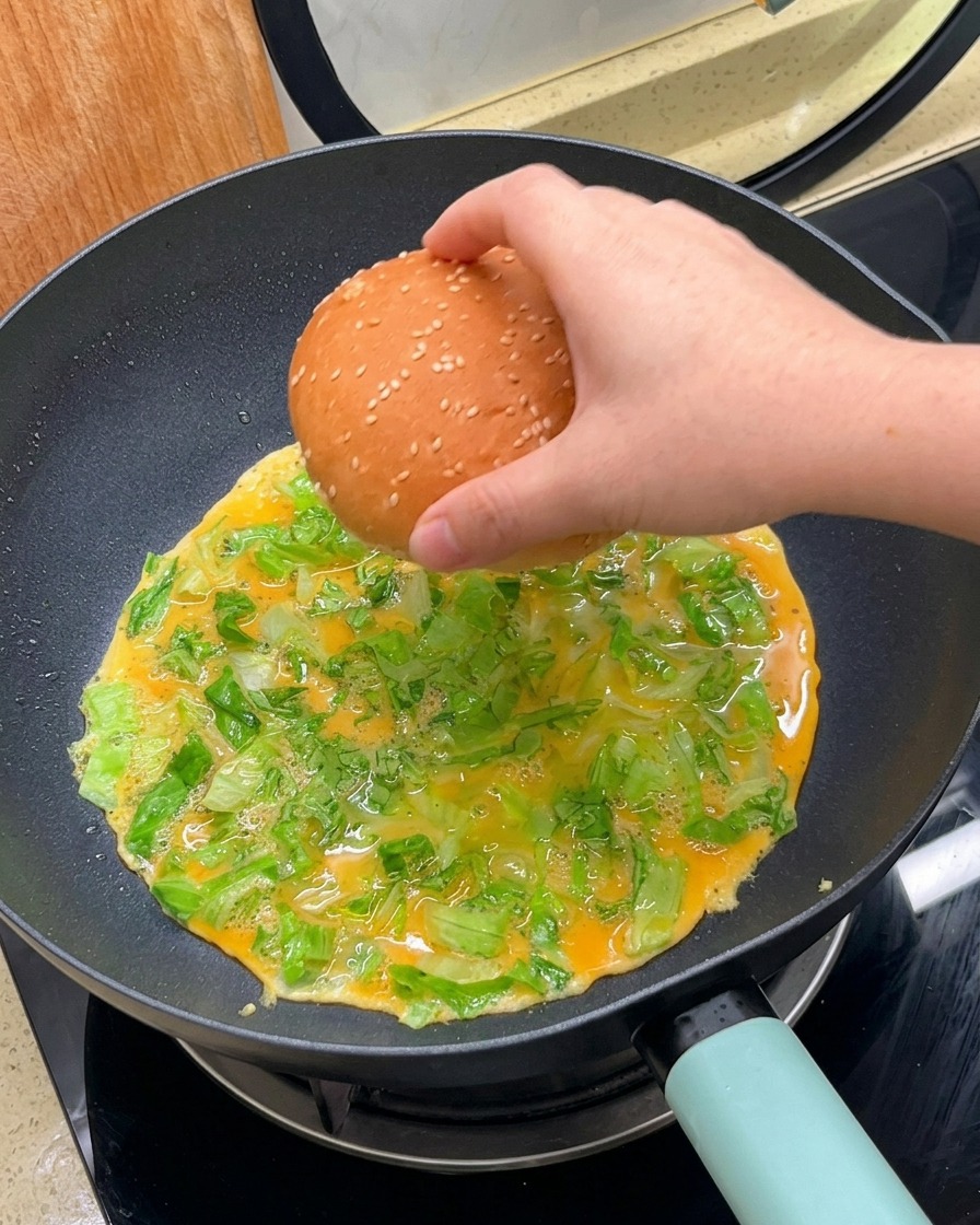 Una mano colocando las dos mitades de un pan de hamburguesa boca abajo sobre la mezcla de huevo y lechuga en la sartén.