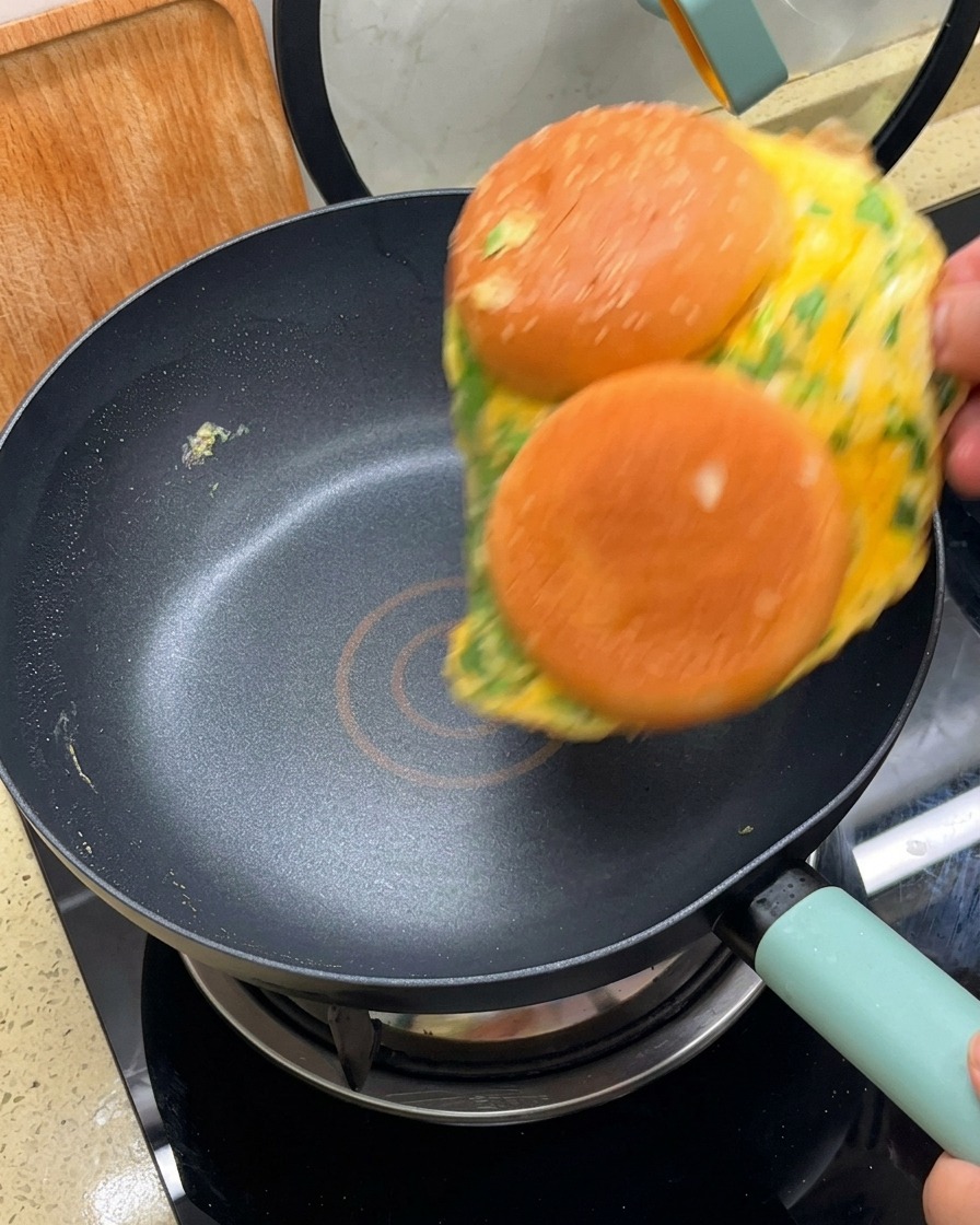 Una tortilla con dos mitades de pan adheridas siendo volteada en una sartén antiadherente negra.