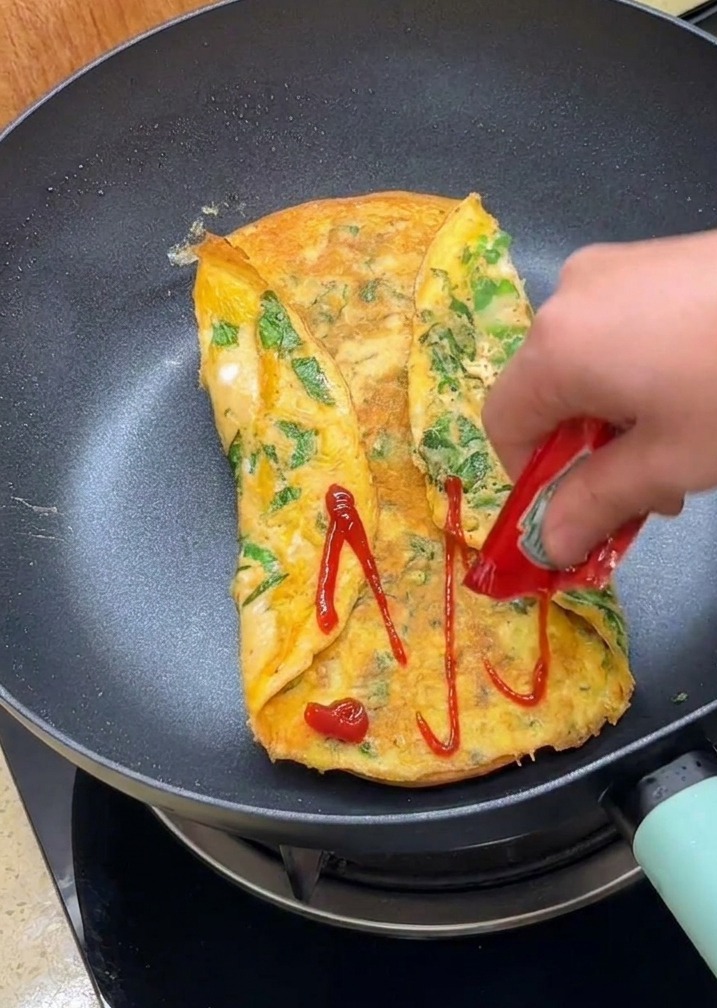 Una mano aplicando kétchup desde un sobre sobre la superficie de la tortilla doblada en la sartén.