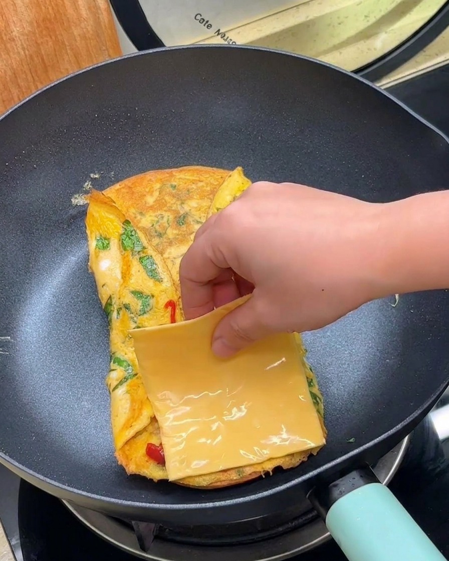 Una mano colocando una loncha cuadrada de queso amarillo sobre el huevo doblado en la sartén.