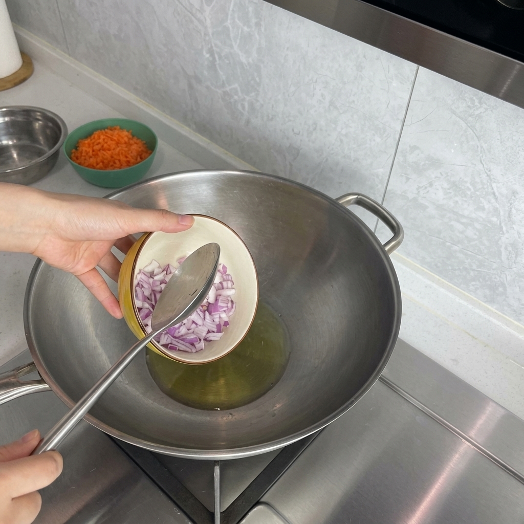 Una mano vertiendo cebolla roja picada desde un bol pequeño a un wok con aceite caliente.