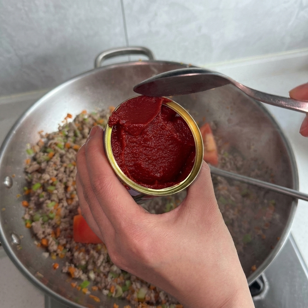 Una persona sosteniendo una lata de pasta de tomate roja y usando una cuchara para echarla al wok con carne y verduras.