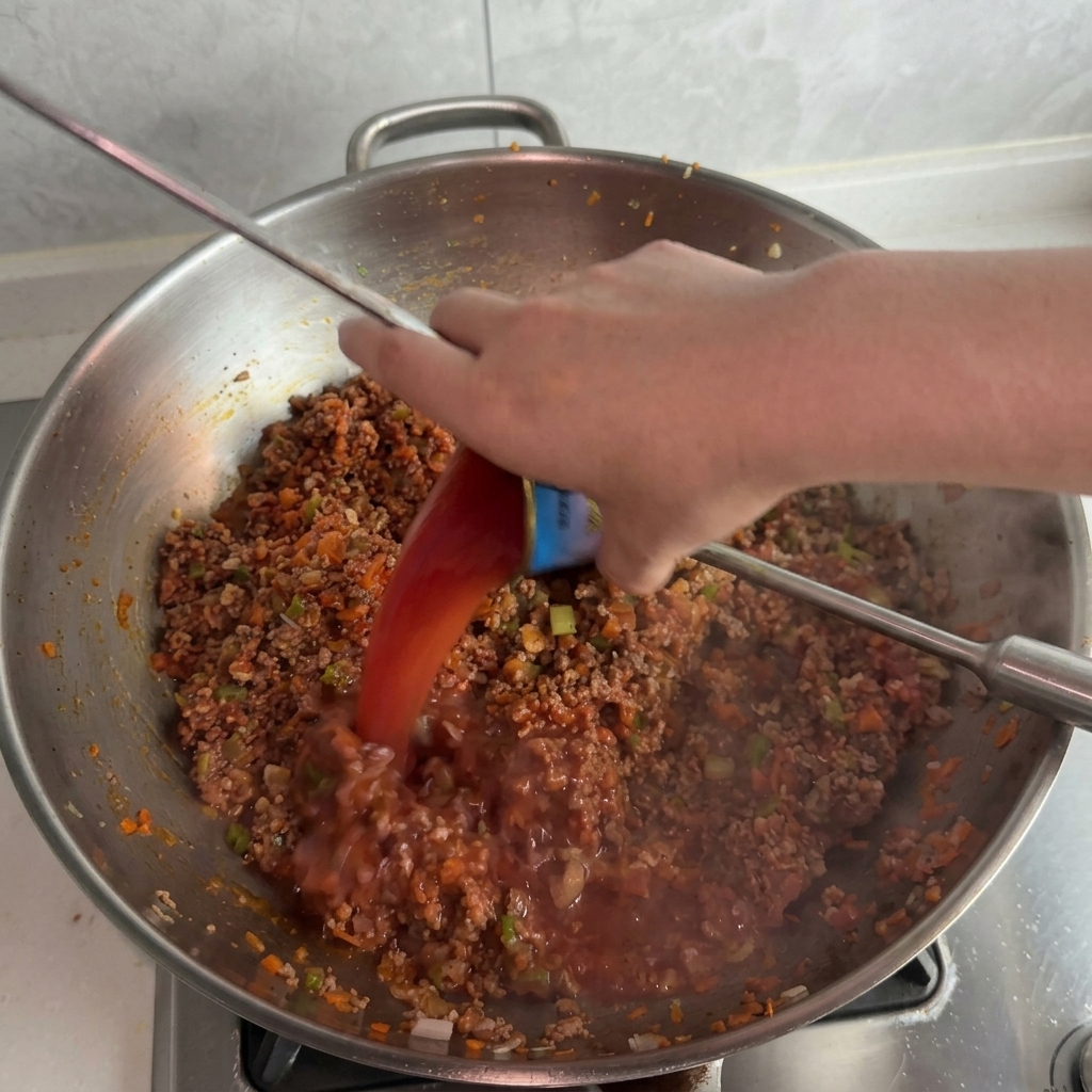 Agua con restos de pasta de tomate vertiéndose desde una lata pequeña a un wok con salsa de carne burbujeante.