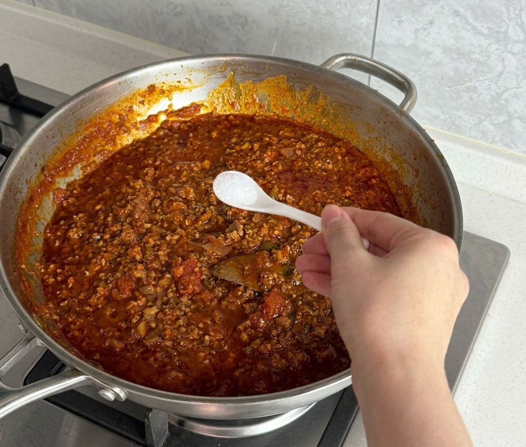 Una mano con una cuchara pequeña añadiendo sal blanca a la salsa de carne roja.
