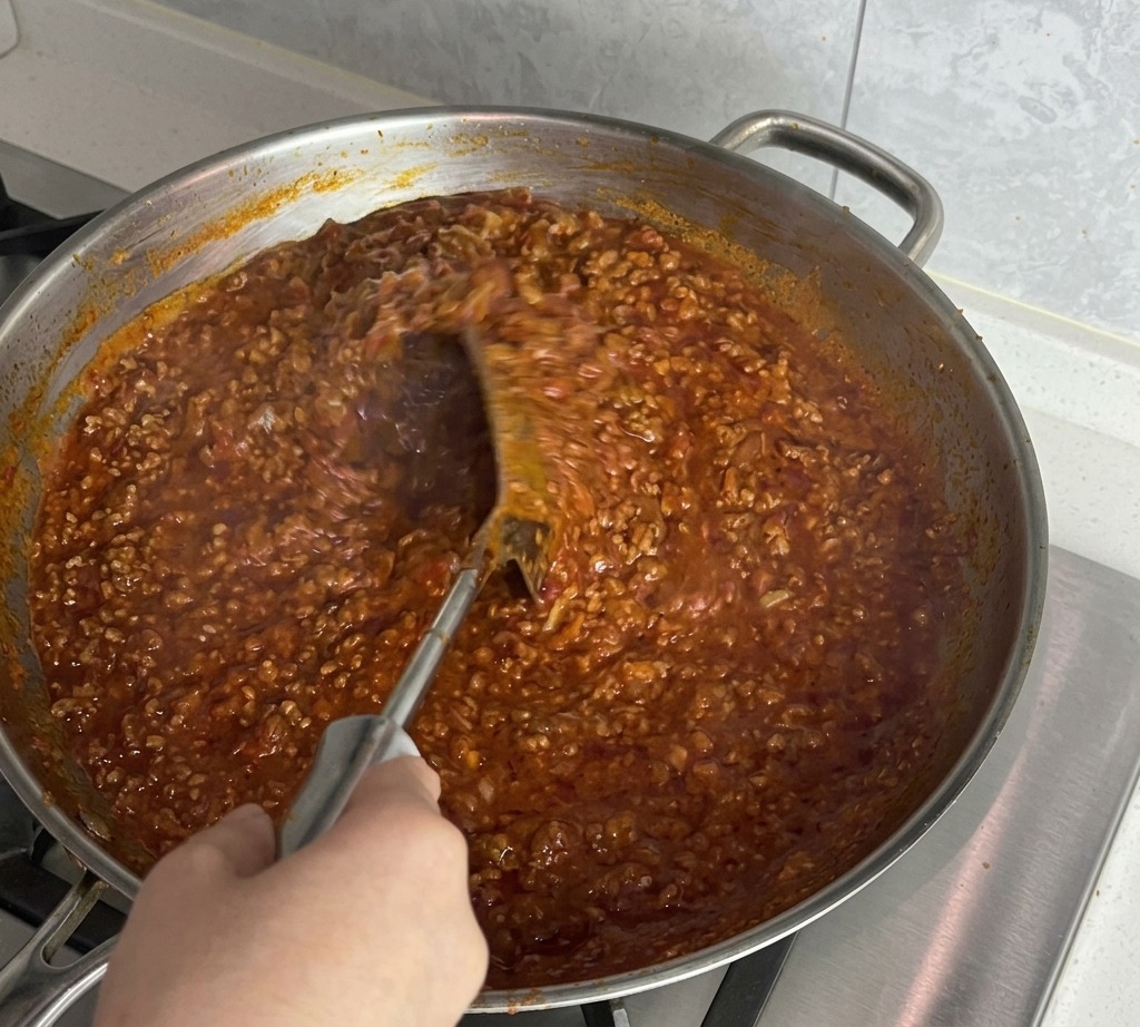 Una mano usando una espátula de metal para remover vigorosamente la salsa espesa de tomate y carne.