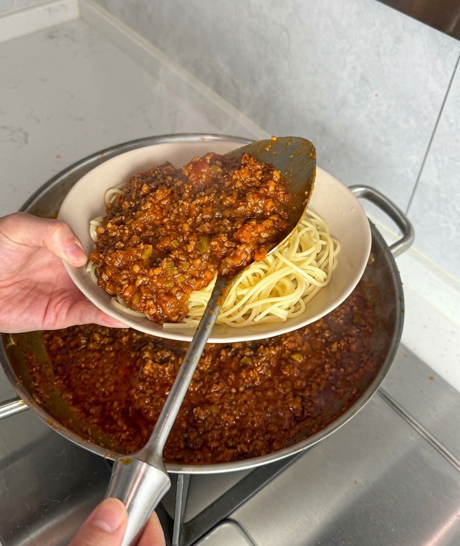 Un cazo sirviendo salsa de carne espesa sobre un plato de espaguetis recién cocidos.