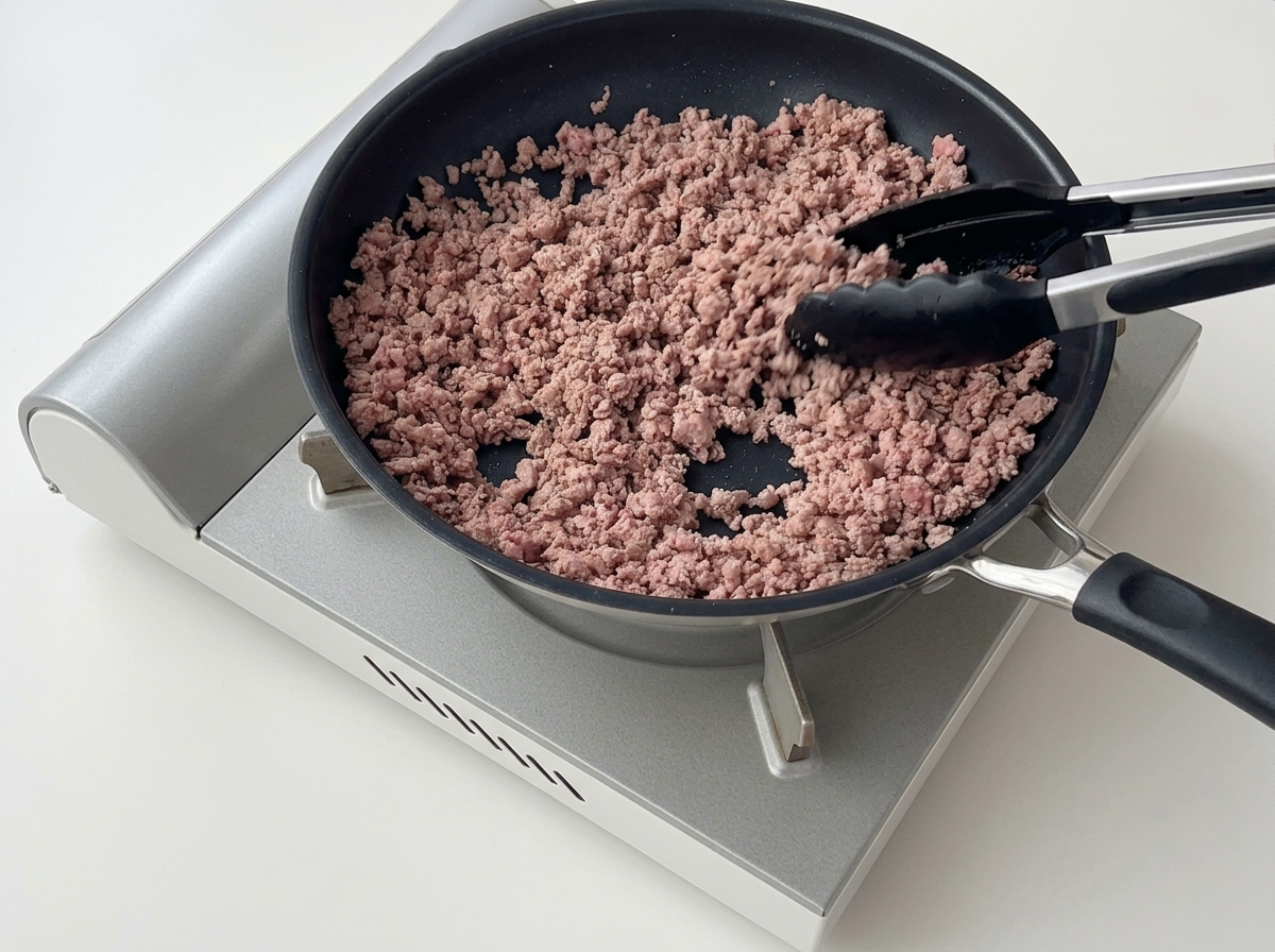 Pinzas desmenuzando y removiendo la carne picada dorada en una sartén antiadherente.