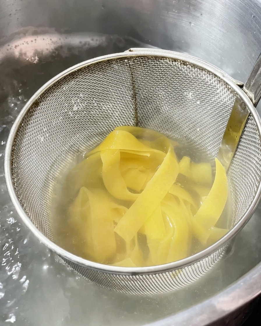 Pasta de cintas anchas cocinándose dentro de un colador metálico sumergido en una olla con agua hirviendo.