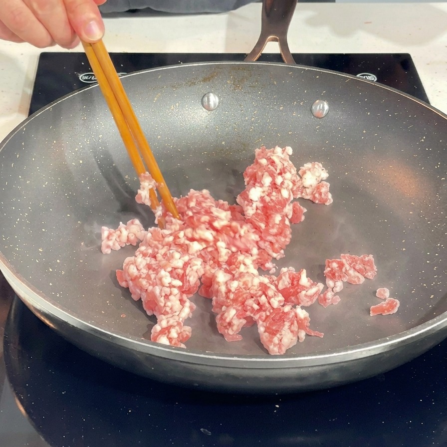 Usando palillos para desmenuzar carne de cerdo picada cruda en una sartén caliente.