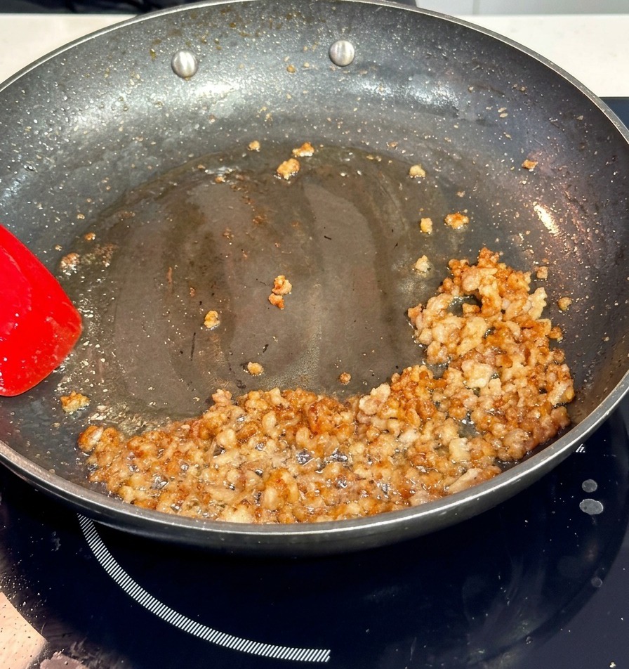 Carne de cerdo picada dorándose en una sartén antiadherente oscura, removida con una espátula roja.