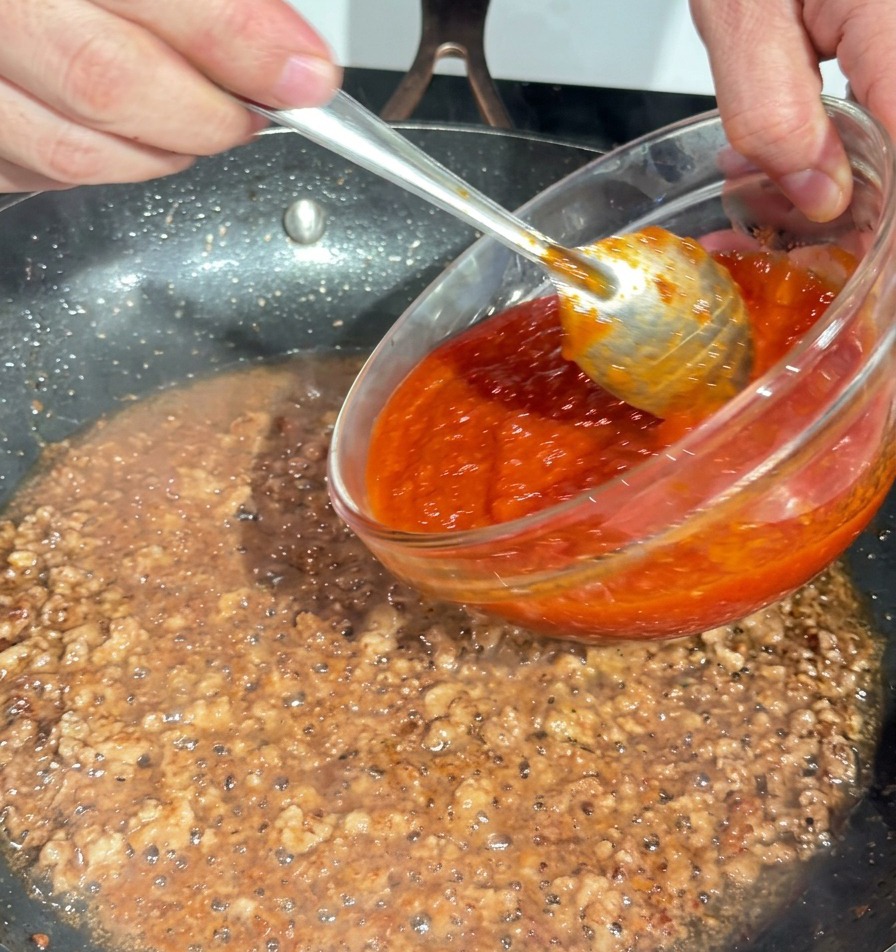 Vertiendo salsa de tomate roja espesa desde un cuenco de cristal a la sartén con la carne dorada.