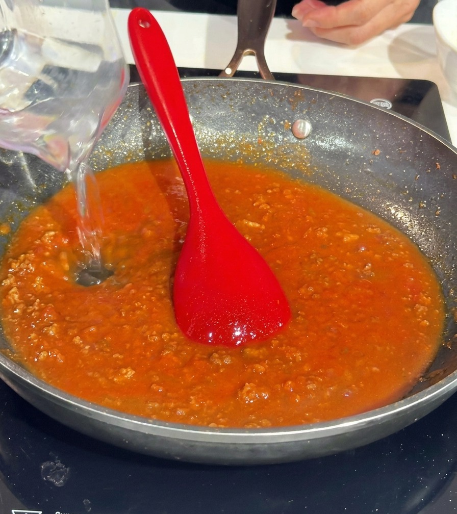 Vertiendo agua desde una jarra medidora transparente a una sartén con salsa boloñesa de tomate.