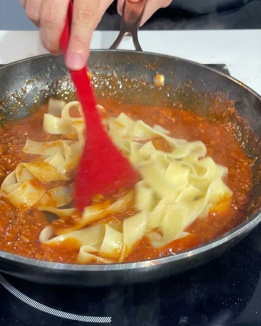 Una espátula roja mezclando las cintas de pasta en la sartén para cubrirlas uniformemente con la salsa boloñesa.