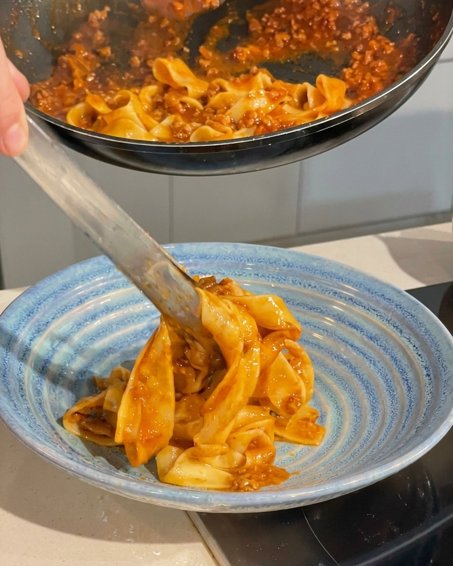 Pinzas metálicas sirviendo pasta con salsa desde la sartén a un plato hondo de cerámica azul.