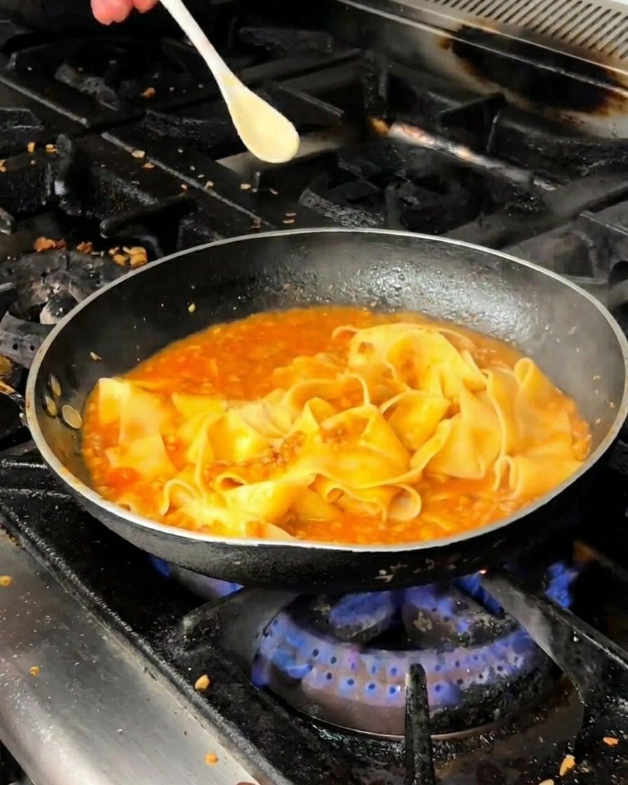 Pasta pappardelle integrándose con la salsa de carne roja en una sartén.