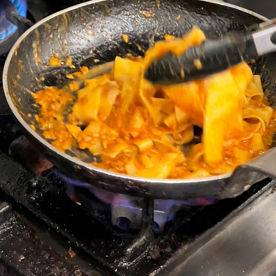 Usando pinzas para mezclar enérgicamente la pasta con la salsa en una sartén.